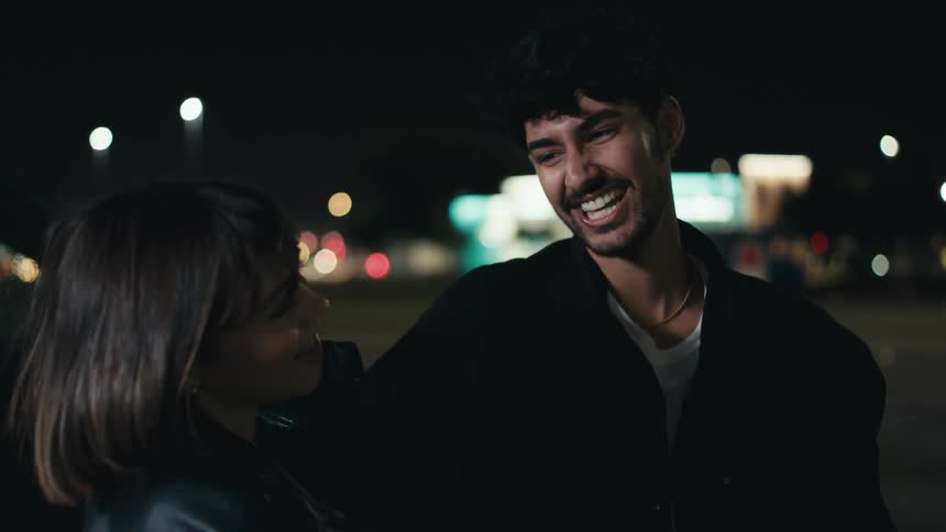 Couple talk and laugh outdoors at night as vehicles pass in the distance