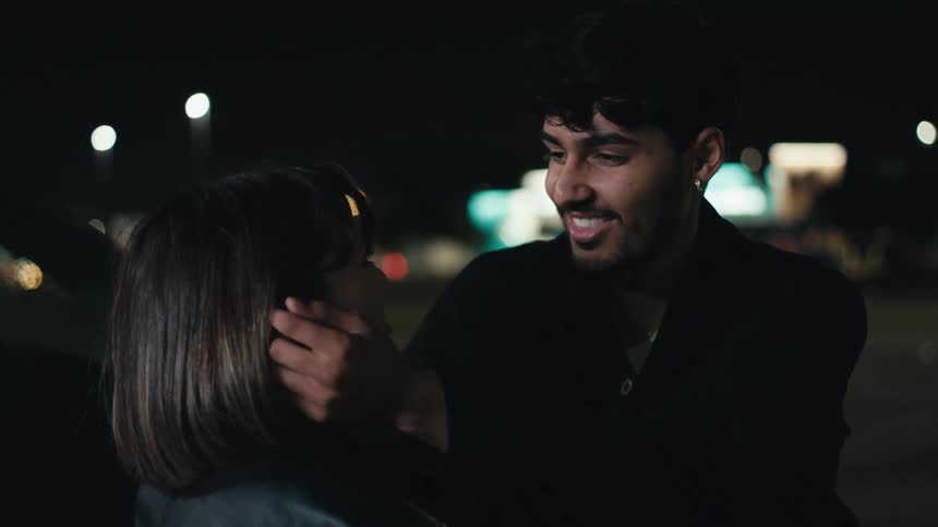 Two lovers smiling and the man passes his hand through the womans hair on a city street at night