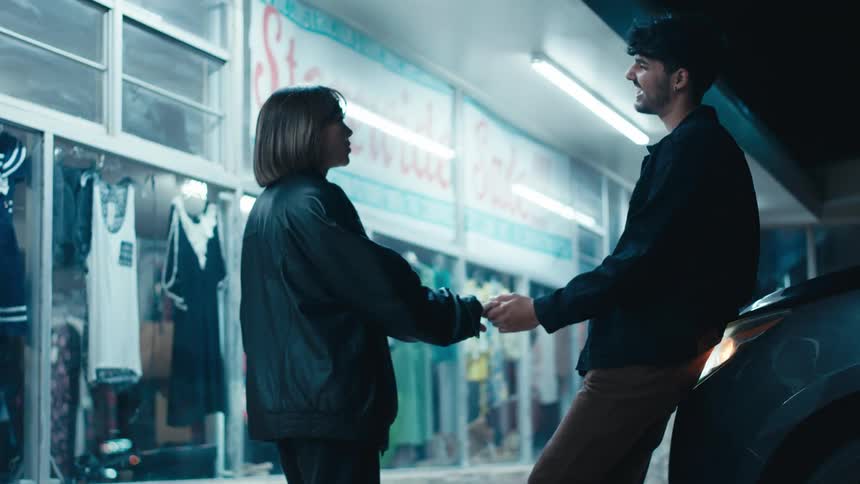 Man leaning against vehicle with its headlights on holds hands with and talks to woman at night