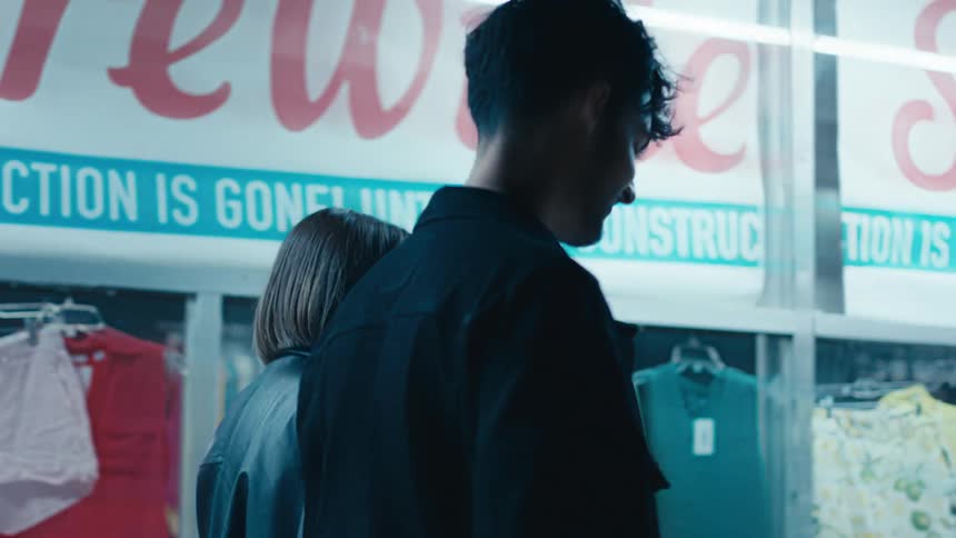 Man and woman walking down a sidewalk in front of a store window at night as they hold hands
