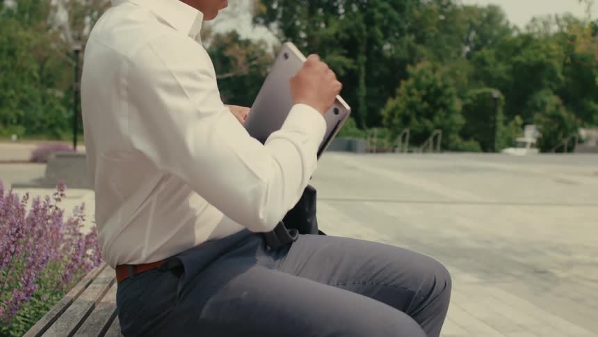 A man sitting on a bench takes a tablet out of his handbag