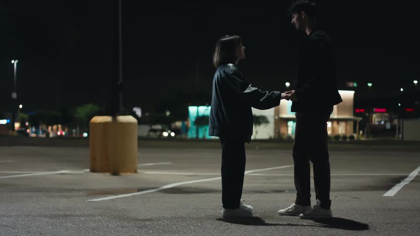 Man and woman in black coats pants and white shoes hold each other on the street at night