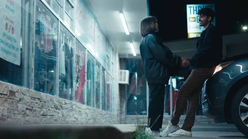 A woman and a man talk and smile while holding hands outside a convenience store as he is leaning on a car