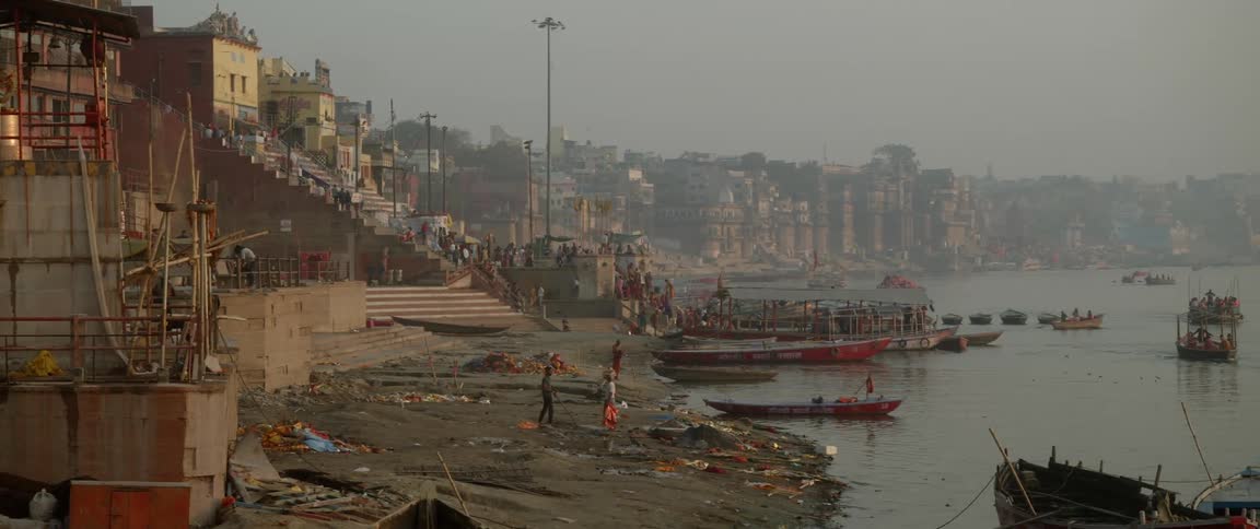 A crowd of people gathered next to a dirt dock while boats sail near it on a dirty river