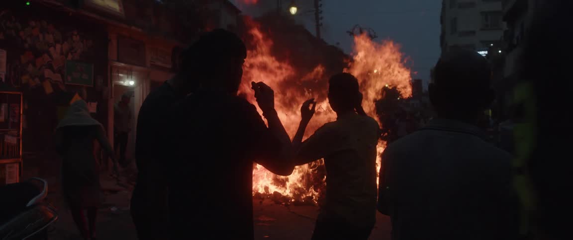People are standing in the street watching a bonfire burn near buildings and one boy is holding a phone up as he walks
