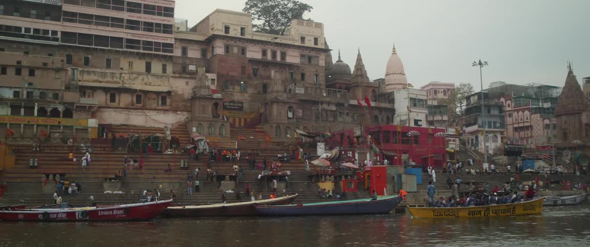 Several people are on canoes on a lake with a neighborhood in the background and many people standing on stairs