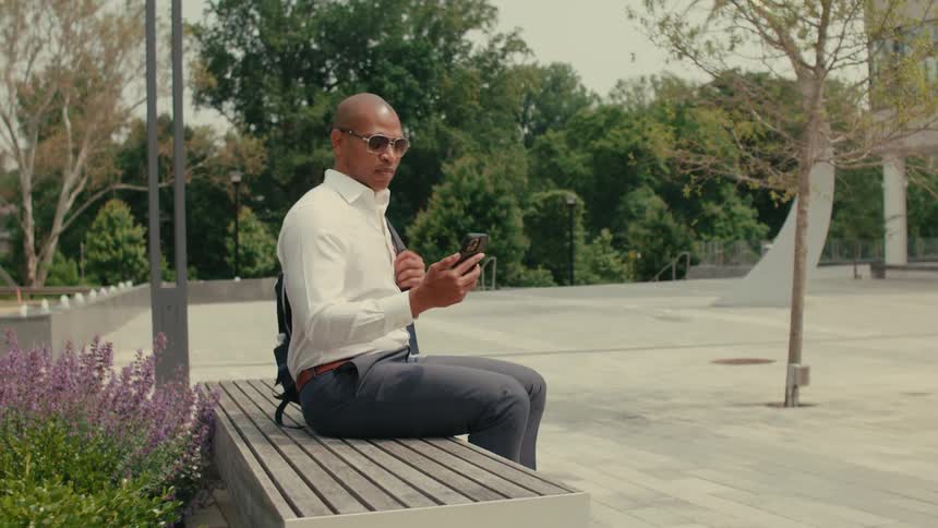 Man carrying backpack and using smartphone set the phone on bench as he sits
