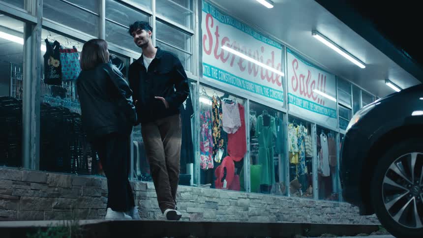 Man with black jacket and brown pants talks with woman wearing black jacket and pants near clothing store