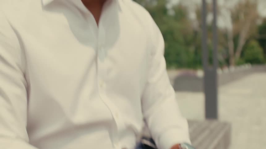 Man sits on bench in park typing on laptop as vehicle passes on road in the background