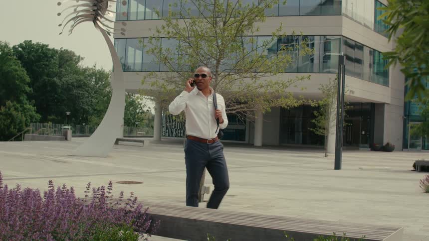 Businessman with sunglasses and white shirt and blue pants talks on smartphone in front of office building