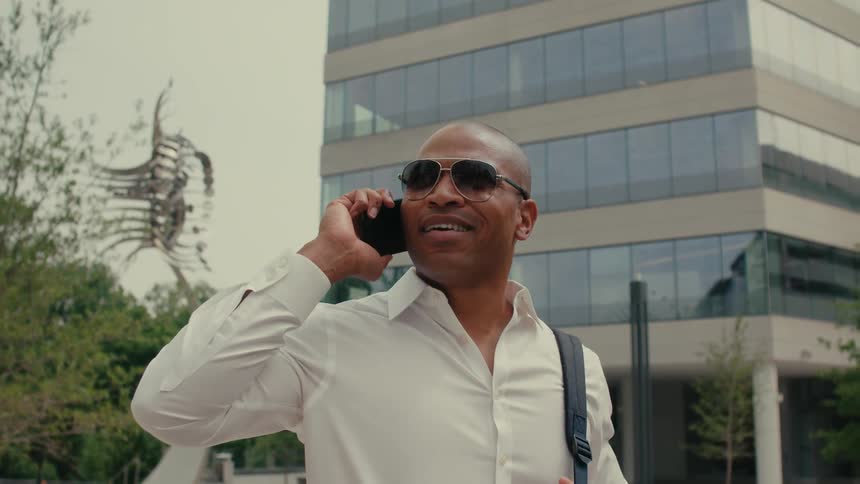 Man wearing dress shirt and sunglasses carrying laptop bag standing on city street in front of building talking on phone