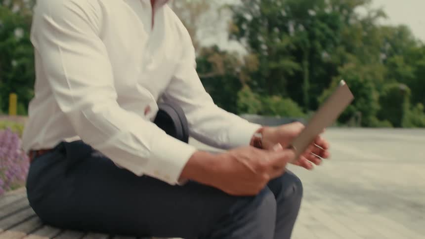 A man is sitting outside on a park bench typing on a laptop computer