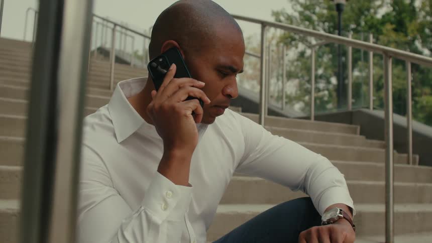 A man is sitting outside on steps near a railing holding his phone to his ear and looking at his wristwatch