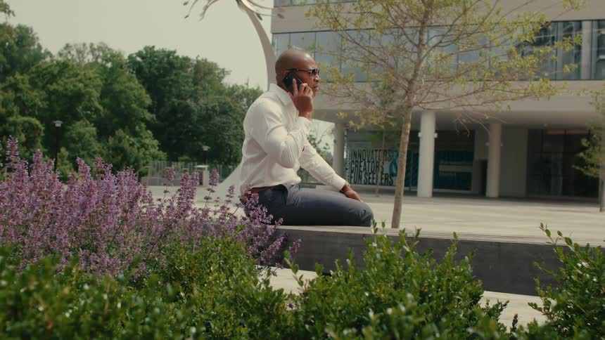 A man sitting down on a concrete bench outside talking on his phone who then interrupts the call to check his phone