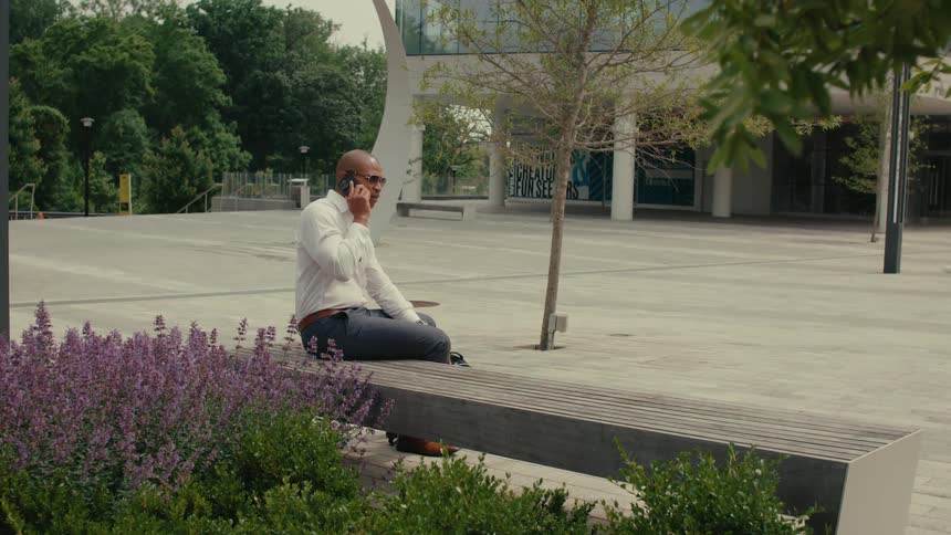Businessman with sunglasses white shirt and blue pants talking on smartphone sits on bench in front of office building