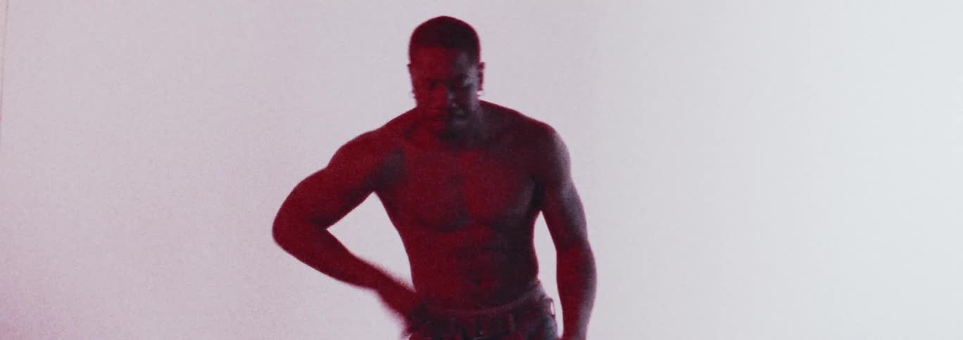 A muscled man dances in a strong determined manner under a red light in front of a white background