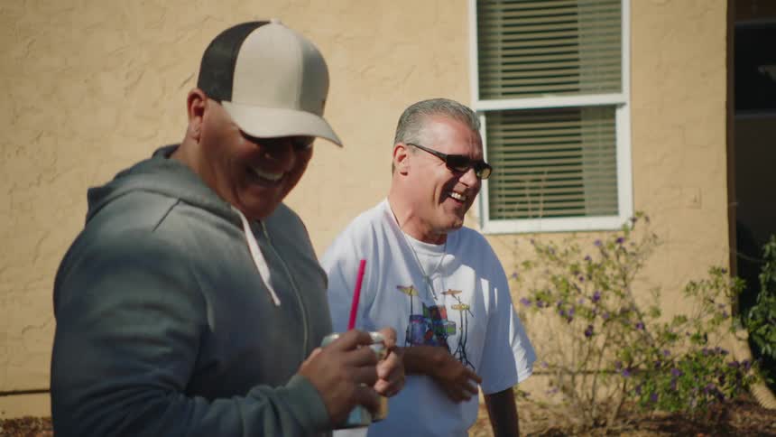 Two men laugh while drinking beer outside of a house