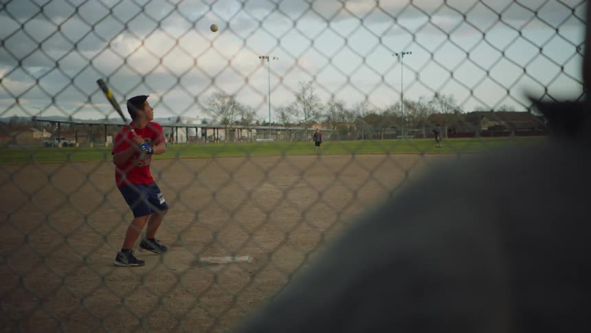 Person stands watching as man tosses up and hits baseball and outfielders wait