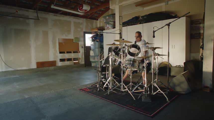 A man sits at a drum kit preparing to play and smiling