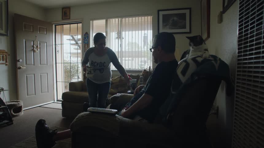 A man with a prosthetic leg is sitting in a lounging chair with a cat behind his head when a woman brings two drinks one for him and one for her that she drinks as she sits on a couch