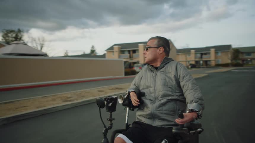 A man in an electric wheelchair is traveling on a neighborhood street looking around as he goes