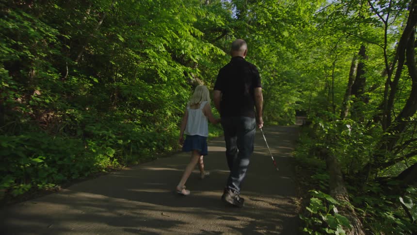 A blind man walks with his granddaughter down shaded path as he uses his walking stick