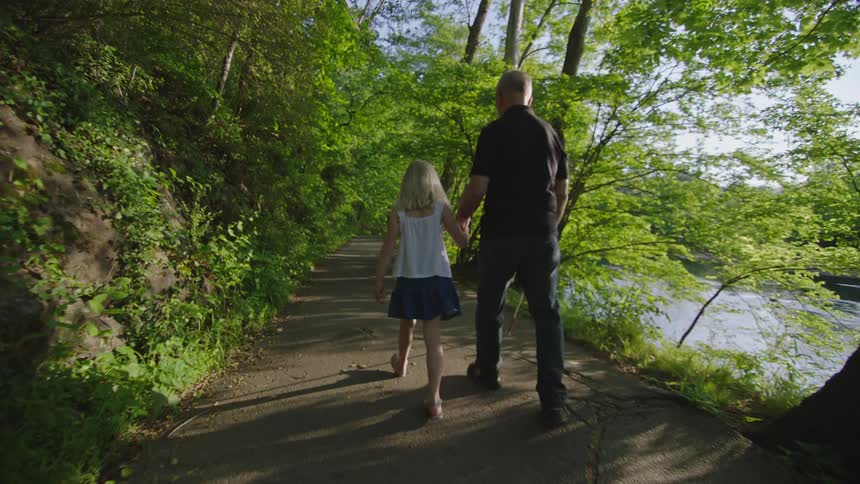 A blind man and his granddaughter walk down a path next to a river