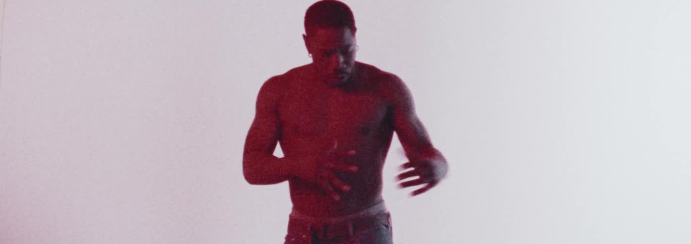 A muscled man bathed in red light dances expressively in front of a white background