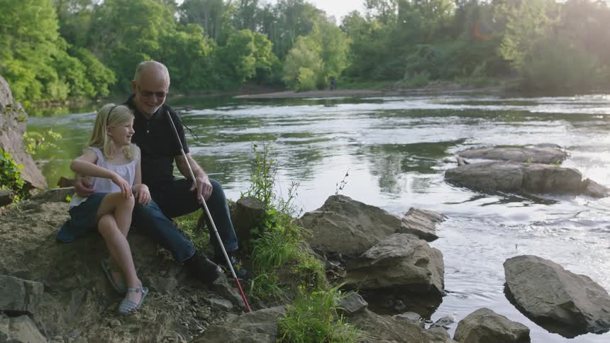 A blind grandfather and his little granddaughter share a laugh as they have a special moment by the river in the summertime