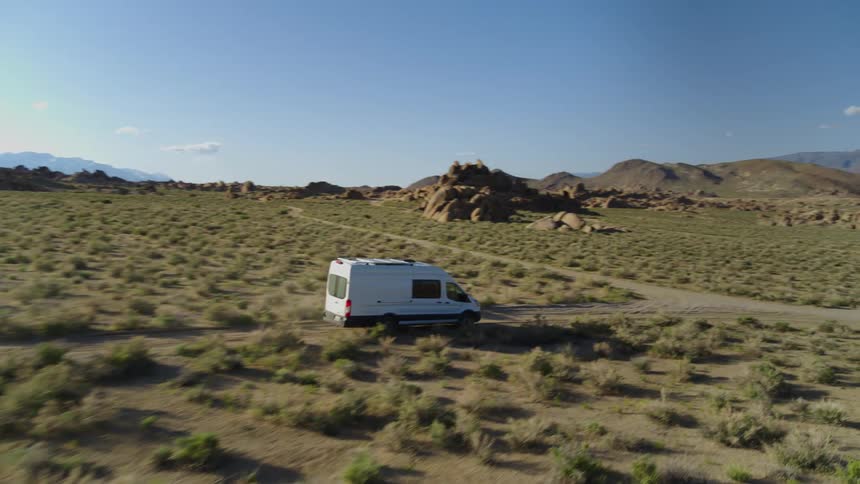 A camper van / delivery truck drives through a dirt road crossing a grassy plain surrounded by rock formations
