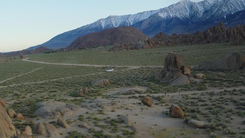 A car drives down a dirt road in the desert with mountains in the background