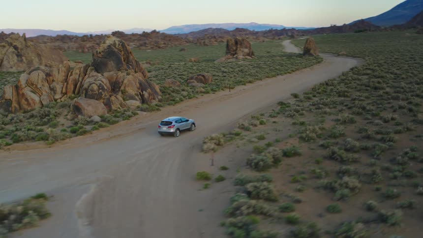 Car drives along dirt roadthrough scrub land passing two intersections with rock outcroppings and mountains in the background