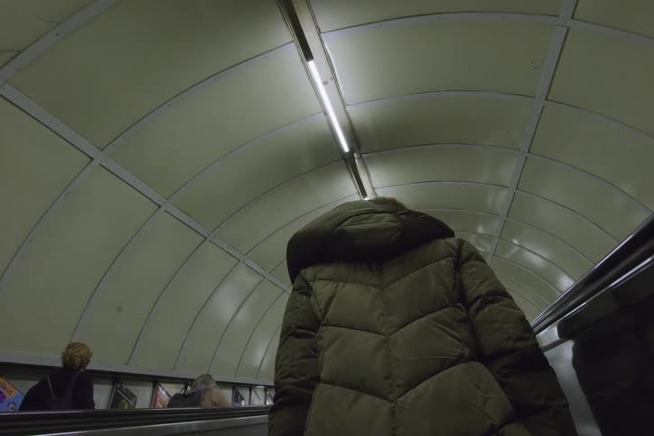 People ride escalator through tube tunnel lit by fluorescent lights