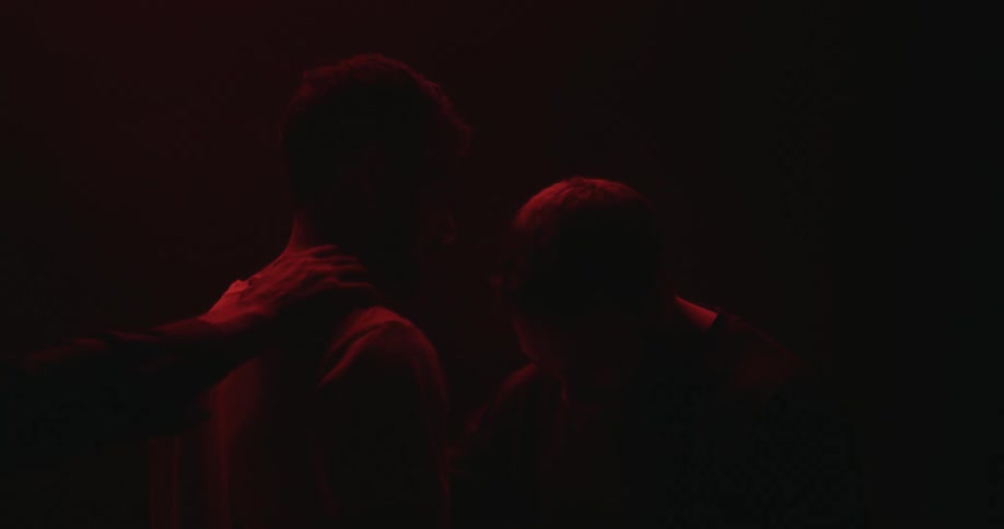 Two man are facing each other in a dark red room one of them with his head bowed when the hand of a third person comes from behind the other man and reaches for his neck