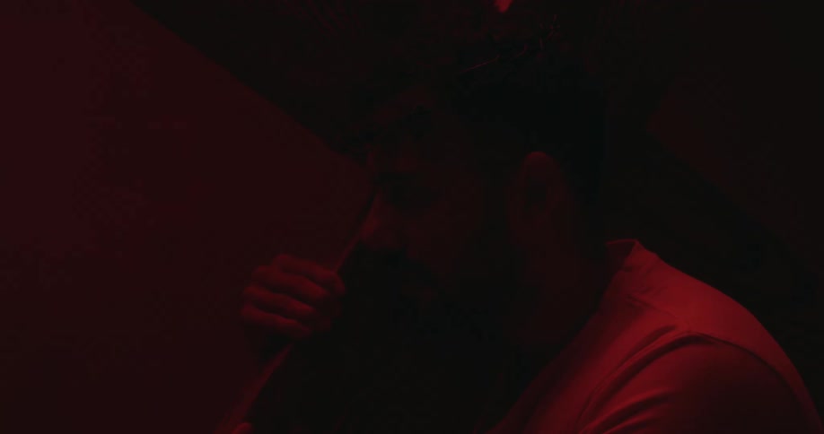 Red light shines on a mans face in the darkness as he holds a piece of wood over his shoulder and looks up