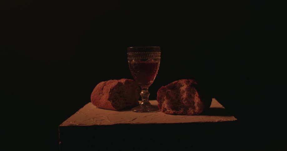 Light shines on a glass goblet filled with beer is sitting on a box with two halves of a loaf of bread in the darkness