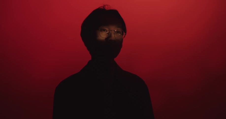 A man in glasses hidden in shadow in front of a red background until light breaks up his silhouette by showing his eyes