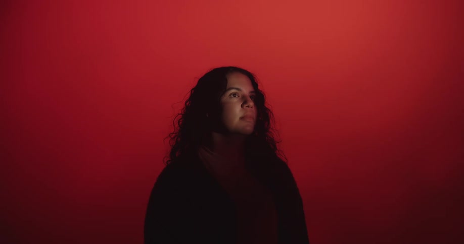 A young hispanic woman stands in a dimly lit area with a red background and a single point light on her face turns head upward into the light and gazes up