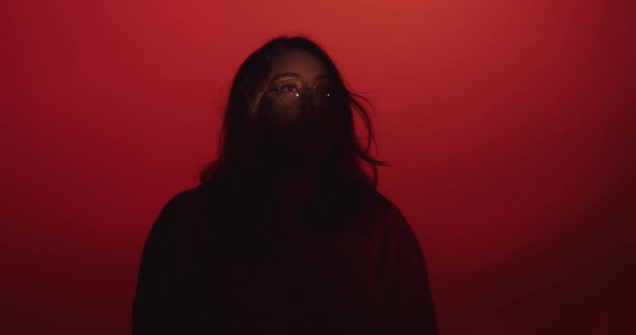 Woman standing in complete shadow in silhouette against a red lit background she looks up toward the distance and her face lands in a single point light showing only one eye and her forehead