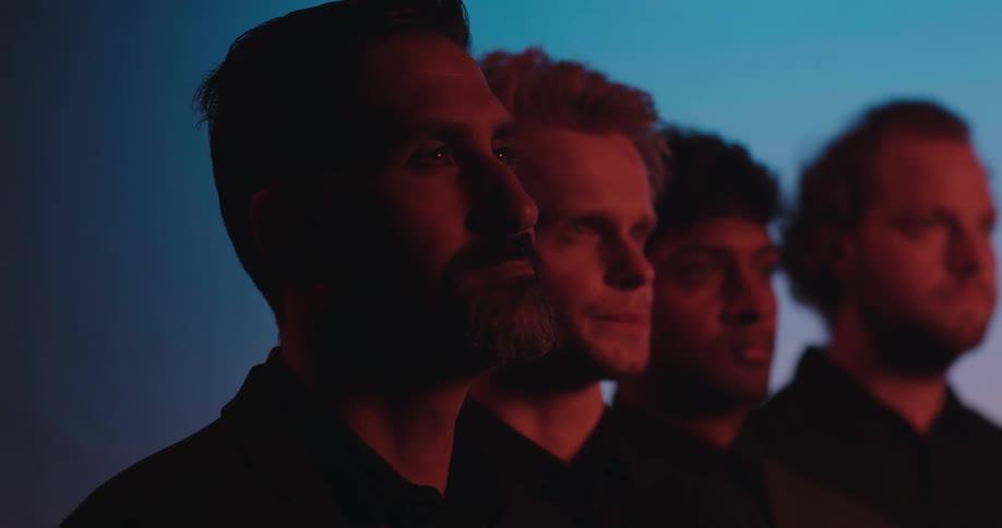 Four men standing side by side against a blue lit background with red light on their faces all three wearing the same black shirt staring off to the right in the distance