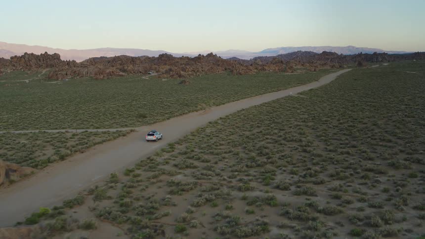 Aerial drone view of white suv turning left on desert rode with mountains in the background late afternoon with the sun illuminating the desert background