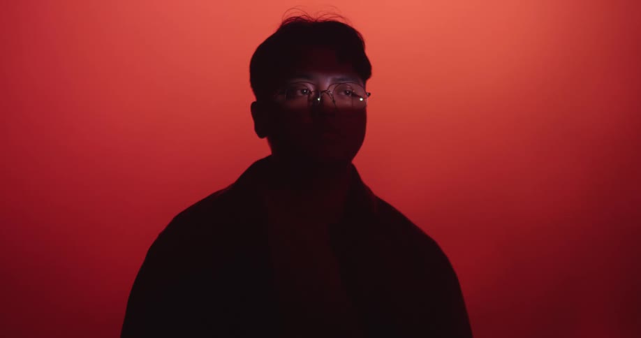 A man in silhouette against a red background with only his eyes and glasses revealed by a ray of light as he turns his head