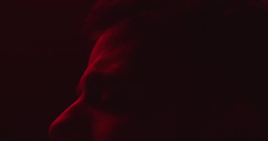 A man lit by red light and in front of a red background is looking up then looks down into shadow looking sad