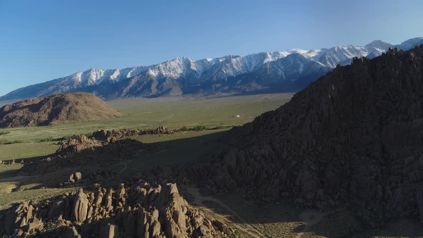 The sun shines brightly upon rock formations and mountains and there are snow covered mountains in the distance under a clear sky