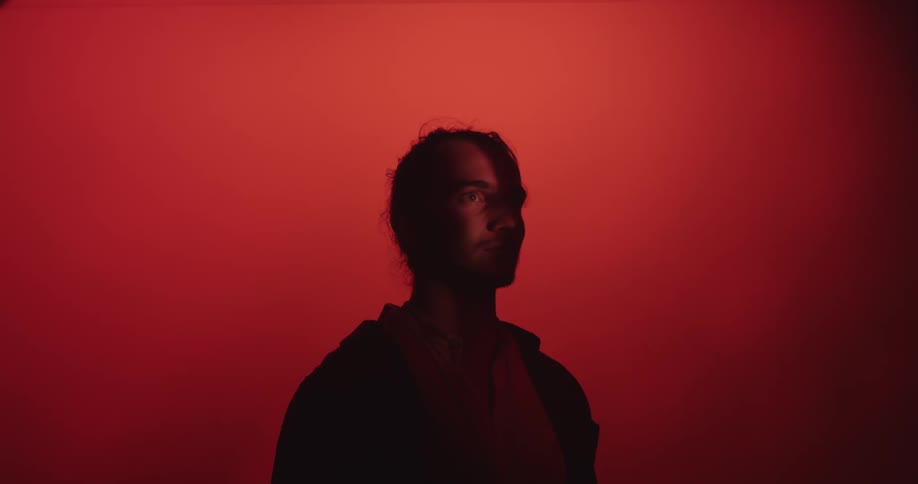 A man is hidden in silhouette in front of a red background until a light shines over his eye