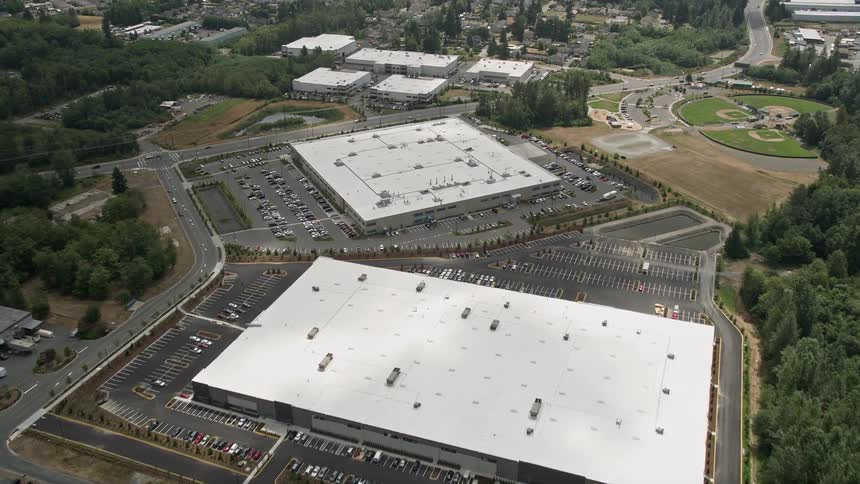 An aerial fly over of a pair of large warehouse buildings in Lynnwood Seattle with a partly cloudy sky and surrounding parking lots filled with cars