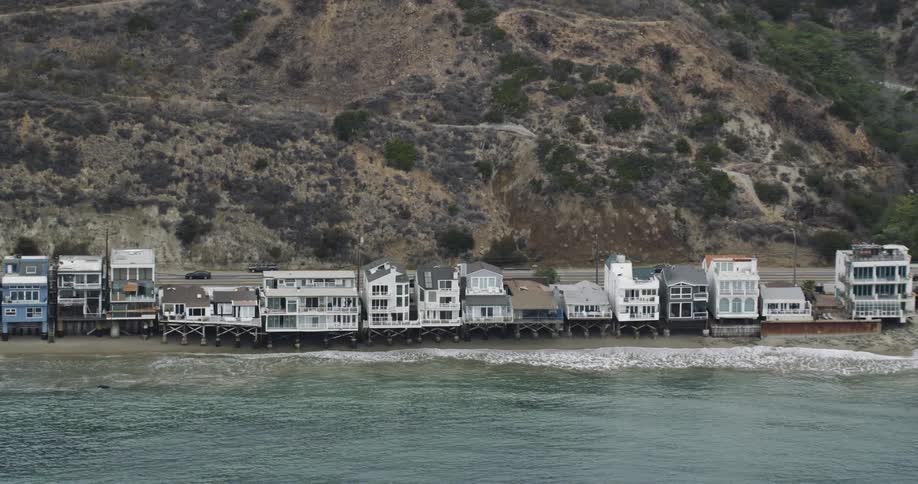 Drone view of houses along a coastline with a road behind the houses with traffic traveling on it at the bottom of a hill