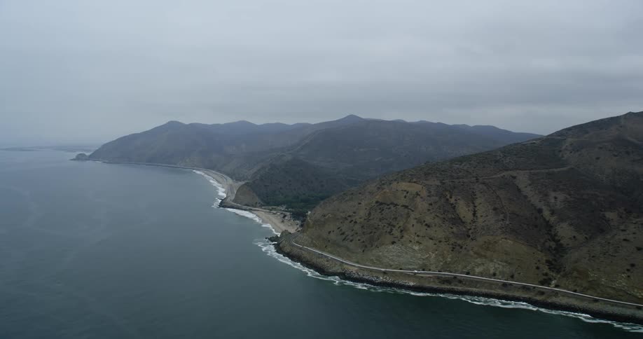 Vehicles move along highway hugging mountains bordering ocean under an overcast sky