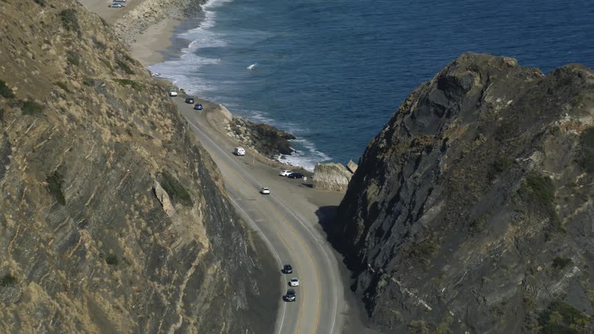 An aerial of a road passing through a mountain while cars travel on it near the coast