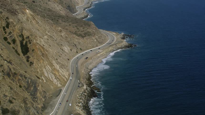 Traffic moves on a mountain road along the coast as waves roll into shore